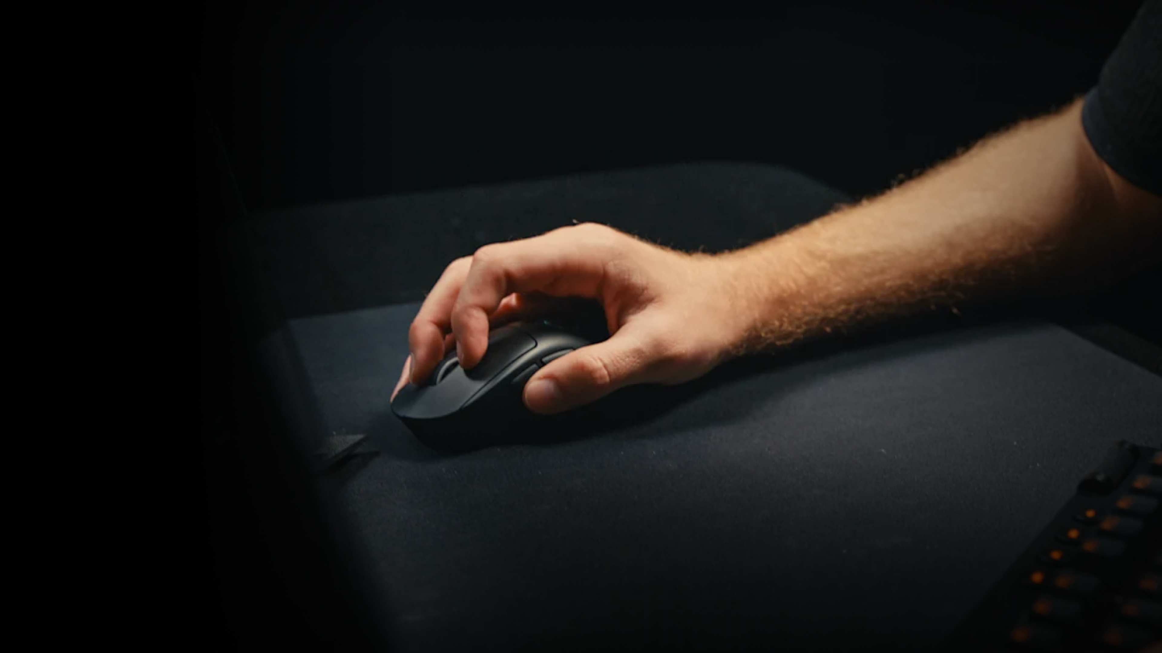 A hand resting on a black Logitech mouse on a dark mouse pad, photographed from the side in low light